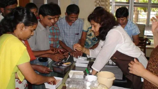 Adults gather around a table, Nita guides them in chocolate preparation.